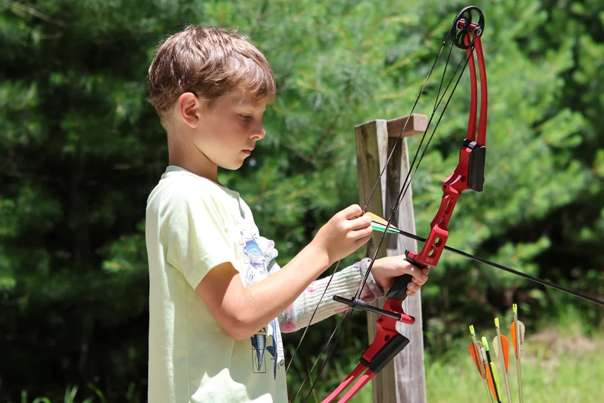 Cub Scout concentrates on preparing an arrow on a red bow at the outdoor archery range.