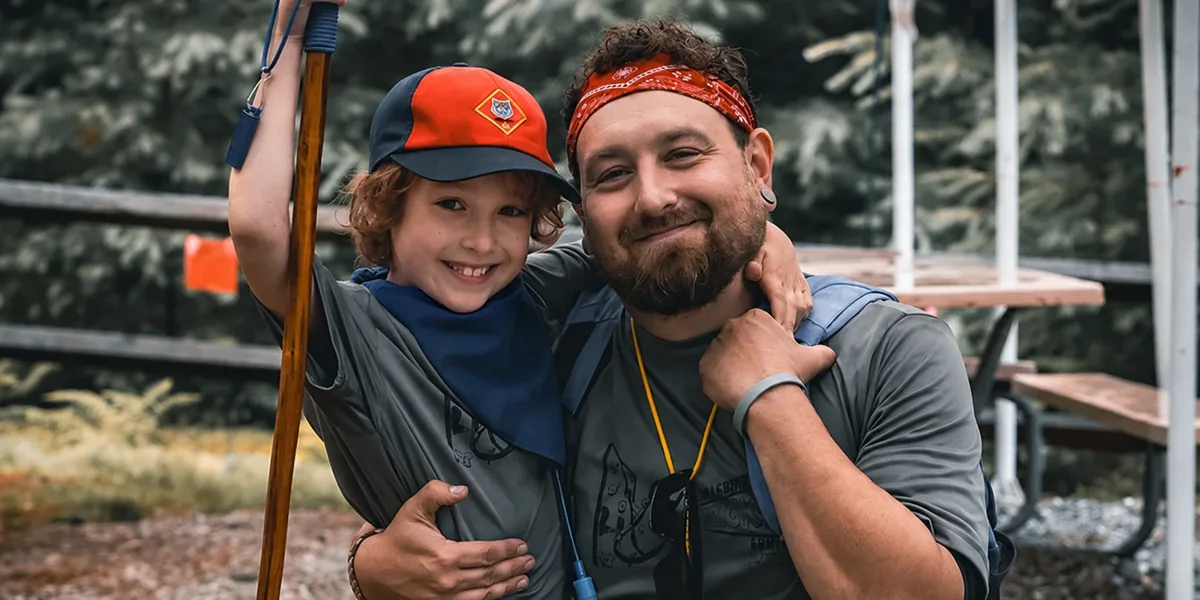 A Cub Scout holding a bow smiles with his father at camp.