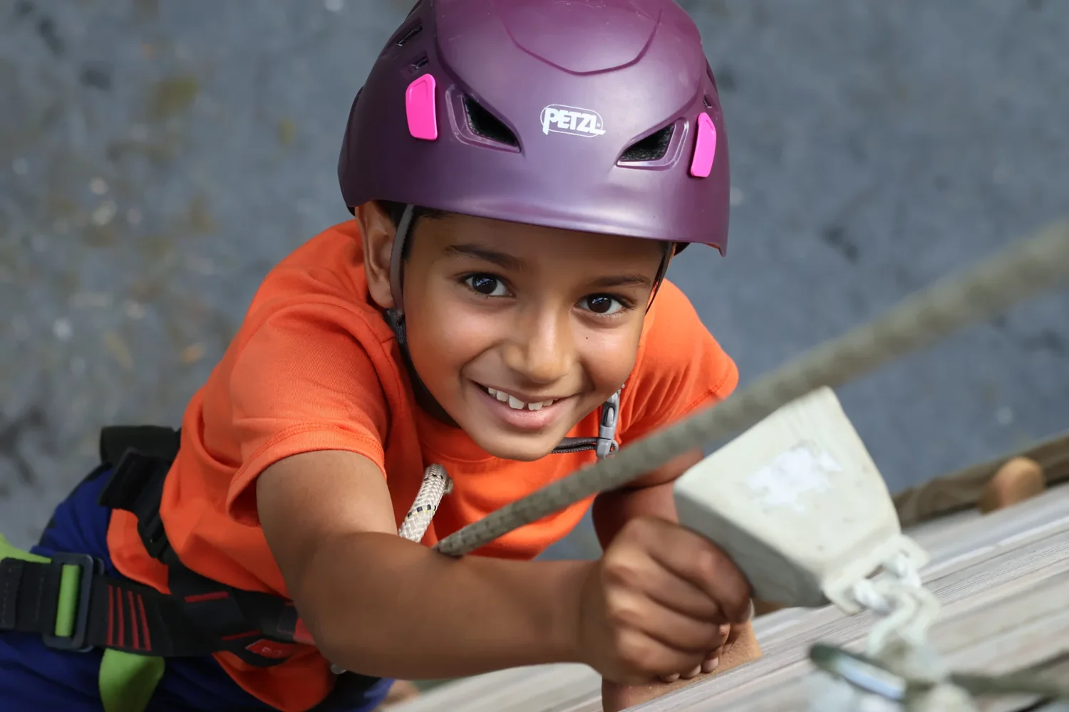 Smiling Cub Scout wearing a helmet and harness reaches up to ring the climbing-wall bell.