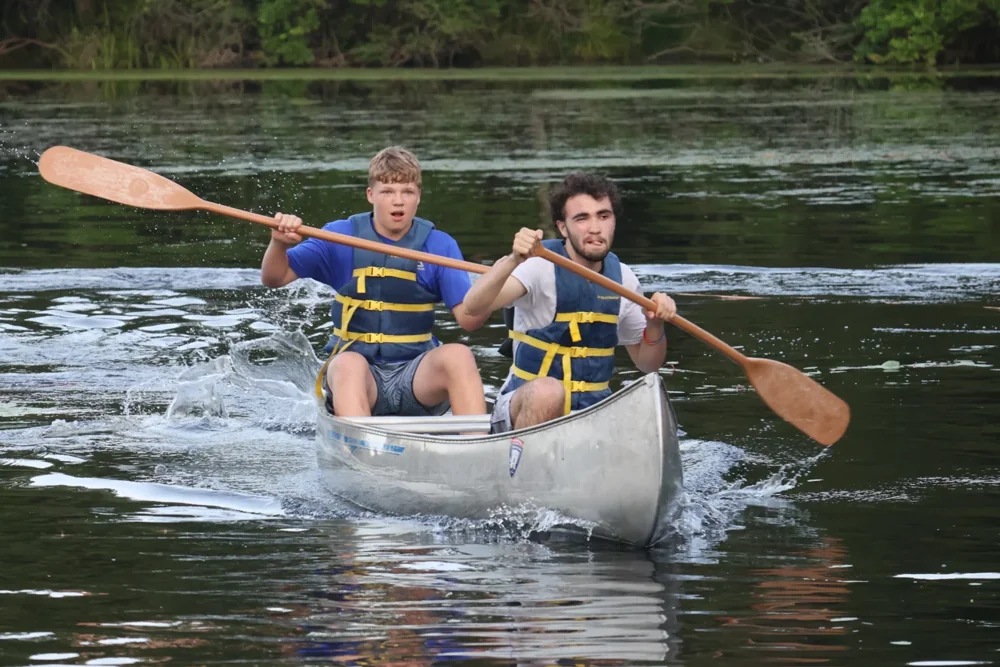 Two Scouts wearing life jackets paddle a canoe across a wooded lake.